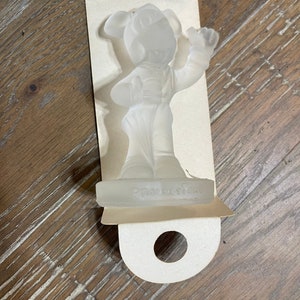 May include: A clear glass figurine of Mickey Mouse in a conductor's uniform. The figurine is standing on a clear glass base with the word "Productions" inscribed on it.