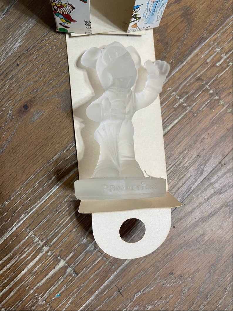 May include: A clear glass figurine of Mickey Mouse in a conductor's uniform. The figurine is standing on a clear glass base with the word "Productions" inscribed on it.