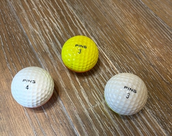 May include: Three white golf balls with black lettering. The balls are labeled "PING 3" and "PING 4". One ball is yellow.