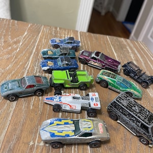 May include: A collection of vintage toy cars in various colors, including green, blue, silver, black, and white. The cars are made of metal and plastic and have a worn, vintage look.