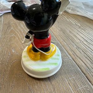 May include: A vintage ceramic Mickey Mouse music box. The figurine is black with yellow shoes and red shorts. The music box base is white with green and yellow stripes.