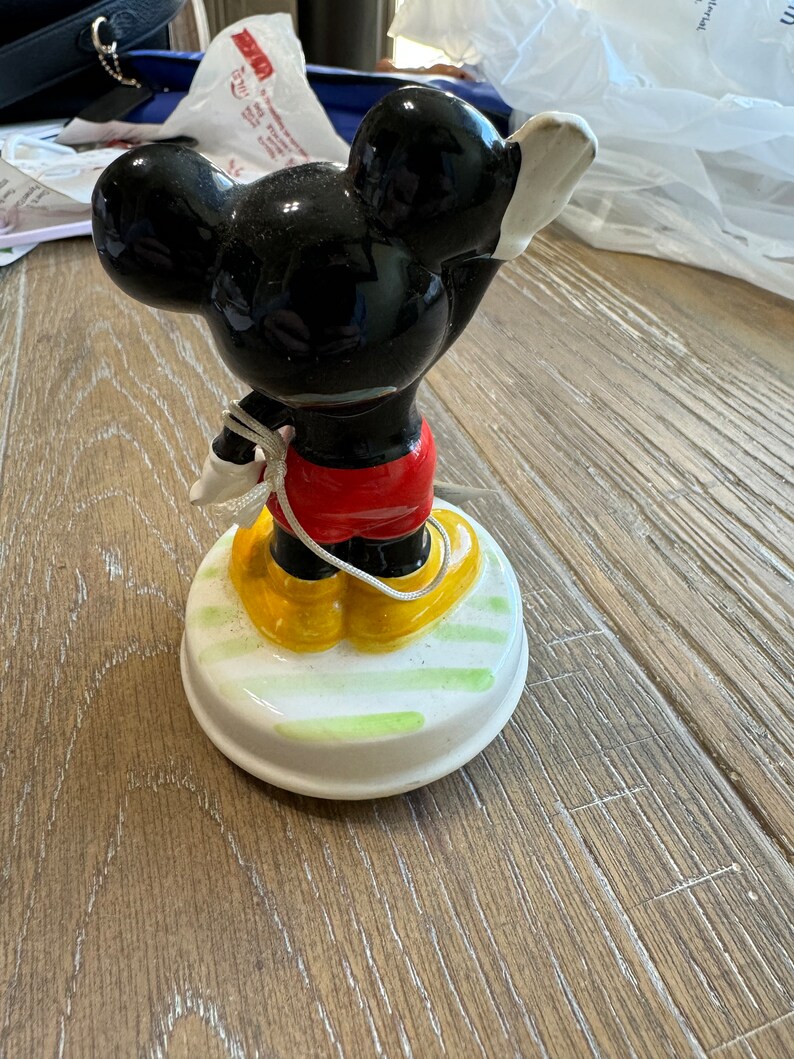 May include: A vintage ceramic Mickey Mouse music box. The figurine is black with yellow shoes and red shorts. The music box base is white with green and yellow stripes.