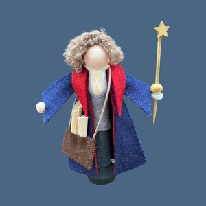 May include: A felt doll wearing a blue robe with a red collar and carrying a brown bag. The doll has brown hair and is holding a gold star-shaped wand.
