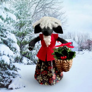 May include: A handcrafted sheep figurine with a black face and ears, wearing a red vest and a floral print skirt. The sheep holds a small woven basket filled with greenery and red berries, tied with a red ribbon. The background shows a snowy winter scene.