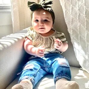 May include: A baby wearing a light brown floral print top with puffed sleeves, blue jeans with distressed details, and a green bow headband.