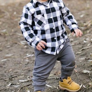 May include: A toddler wearing a black and white plaid button-up shirt with a grey pair of pants and yellow shoes.
