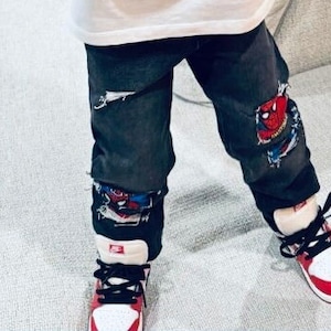 May include: A child wearing black jeans with red and blue Spiderman patches and red and white sneakers.
