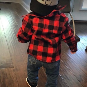 May include: A toddler wearing a red and black plaid flannel shirt, blue jeans, and a black baseball cap with a white arrow and finger pointing up design.