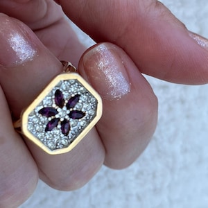 May include: A gold ring featuring a rectangular face with a cluster of six marquise-cut deep purple stones arranged in a flower pattern, surrounded by a pavé of small clear stones set against a silver-toned background within a gold border.