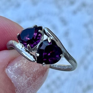 May include: A silver ring featuring two heart-shaped, deep purple faceted gemstones set side-by-side in a bypass style band. The metal band has a smooth, polished finish and the stones are secured with small metal prongs.