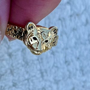 May include: A gold-toned metal ring featuring a sculpted tiger head centerpiece with black enamel detailing on the eyes and ears. The band of the ring has a textured, irregular surface pattern that extends from the tiger head around the finger.