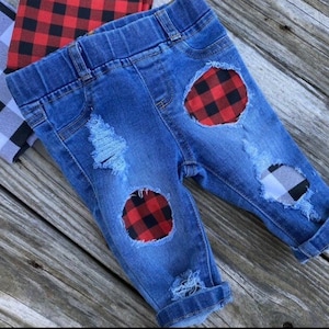 May include: Distressed denim jeans with red and black plaid fabric patches. The jeans have a cuffed hem and are made for a baby or toddler.