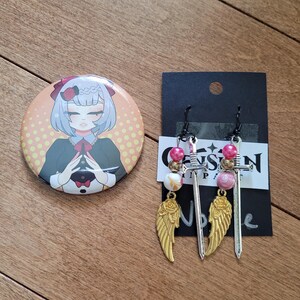 Puede incluir: Un botón redondo con una ilustración de dibujos animados de un personaje con cabello azul y un atuendo verde oscuro. Junto a él, dos pendientes con colgantes de espada plateados, cuentas rosas y blancas y colgantes de alas doradas.