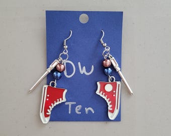 Op de afbeelding: Handgemaakte oorbellen met rode sneakerbedels en witte rand. De oorbellen hebben zilveren haken, kralen en een zilveren naaldvormige bedel. De oorbellen worden getoond op een blauwe kaart met de handgeschreven tekst "Dw Ten".