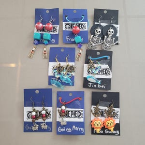 Puede incluir: Una colección de pendientes hechos a mano exhibidos en tarjetas azules y negras. Los pendientes presentan varios diseños, incluyendo esqueletos guitarristas, leones de dibujos animados y temas náuticos. Algunas tarjetas tienen texto como "Going Merry" y "Jin bei".