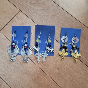 May include: Three pairs of handmade earrings on blue cards. The earrings feature various designs, including silver axes, eagle wings, swords, and dragons. Some earrings have colorful beads and decorative elements.