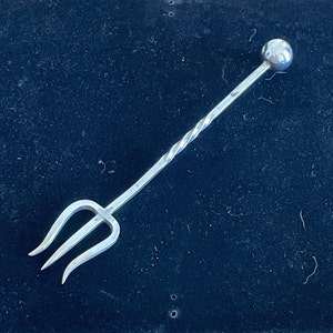 May include: A silver cocktail muddler with a three-pronged fork at one end and a spherical ball at the other. The central shaft has a twisted design. The muddler is made of metal and is set against a dark background.