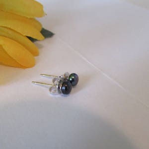 May include: A pair of small, dark gray pearl stud earrings with silver posts. The earrings are set against a white background, with a yellow sunflower in the upper left corner.