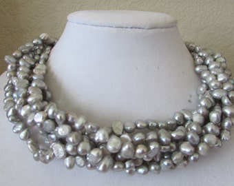 May include: A multi-strand necklace made of grey baroque pearls. The pearls are irregular in shape and size, and they are strung together in a loose, flowing style.