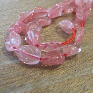 May include: A strand of pink rose quartz beads with a carved leaf design. The beads are strung on a red thread.