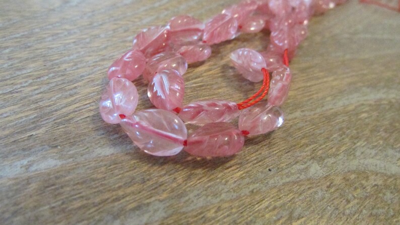 May include: A strand of pink rose quartz beads with a carved leaf design. The beads are strung on a red thread.