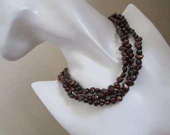 May include: A three-strand necklace made of dark brown baroque pearls. The pearls are irregular in shape and have a slightly iridescent sheen.
