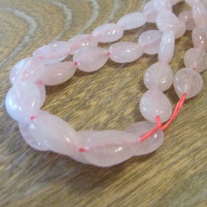 May include: A strand of pale pink oval-shaped beads strung on a red thread. The beads are smooth and translucent.