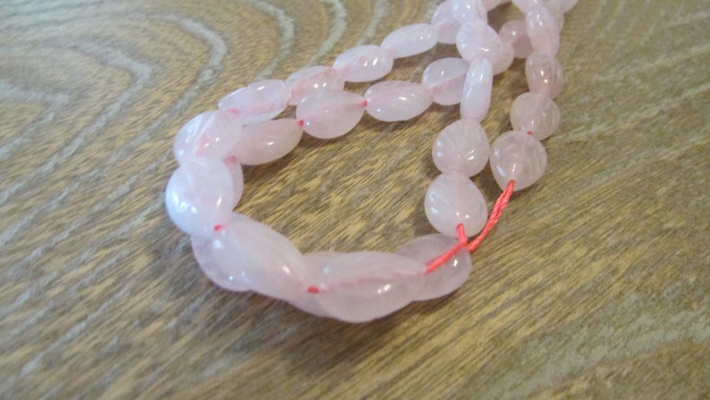 May include: A strand of pale pink oval-shaped beads strung on a red thread. The beads are smooth and translucent.