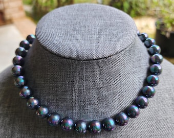May include: A dark iridescent pearl necklace displayed on a grey jewellery bust. The pearls are round and have a lustrous sheen, with hints of blue, green, and purple. The necklace is a classic jewellery piece.