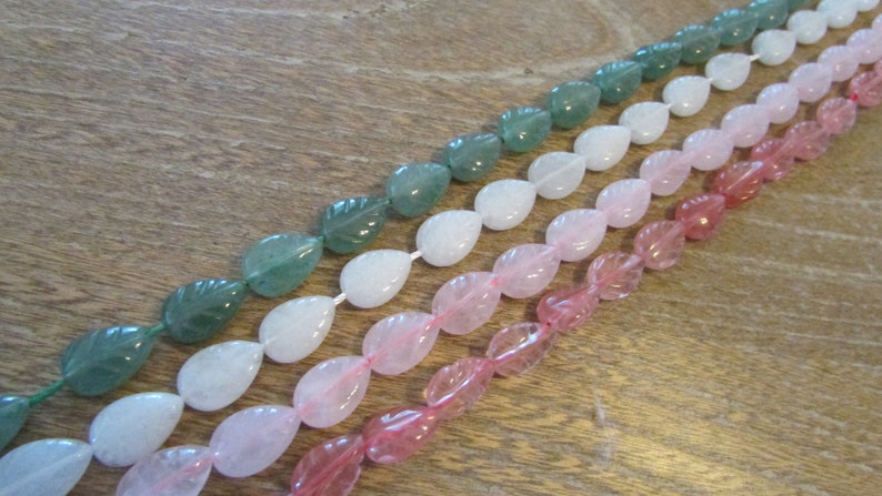May include: A strand of gemstone beads in three colours: green, white, and pink. The beads are shaped like teardrops with a leaf design carved into them.