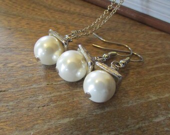 May include: A jewelry set featuring a necklace and earrings. The set includes a silver-toned chain necklace and fishhook earrings, each adorned with a large, round, white faux pearl. The pearls are attached to silver-toned metal accents.