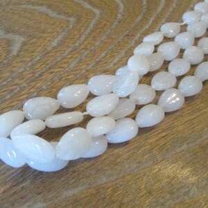 May include: A strand of white teardrop-shaped gemstone beads. The beads are smooth and polished, and they are strung on a white thread.
