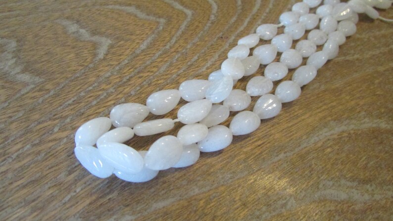 May include: A strand of white teardrop-shaped gemstone beads. The beads are smooth and polished, and they are strung on a white thread.