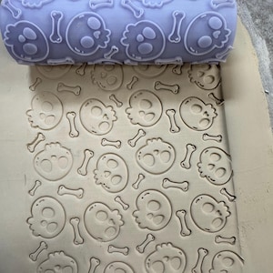 May include: A light purple rolling pin with a skull and crossbones pattern is shown next to a clay surface with the same embossed design. The rolling pin is used to create the skull and crossbones pattern on the clay surface.
