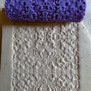 May include: A purple, floral-patterned rolling pin rests atop a sheet of clay, which has been imprinted with the same design. The embossed pattern features ornate scrollwork and floral elements, creating a detailed, textured surface.