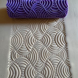 May include: A purple patterned rolling pin next to a textured surface with a matching design. The rolling pin has a repeating, curved line pattern. The surface shows the embossed design, creating a tactile, repeating pattern.