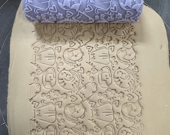 May include: A light purple rolling pin with a cat and heart pattern is pressed into clay, creating an embossed design. The pattern includes cartoon cats, hearts, and paw prints. This tool is for crafting and creating decorative textures.