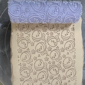 May include: A lavender rolling pin with a bee and flower pattern is pressed into clay, creating an embossed design. The clay is a light tan color, and the rolling pin is made of a hard plastic material. The design includes bees and flowers.