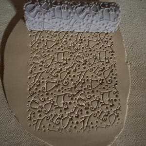 May include: A white rolling pin with a repeating cat design is pressed into a piece of clay, creating a textured pattern. The clay is light brown, and the design includes cats, dots, and swirls. The rolling pin is at the top of the image.