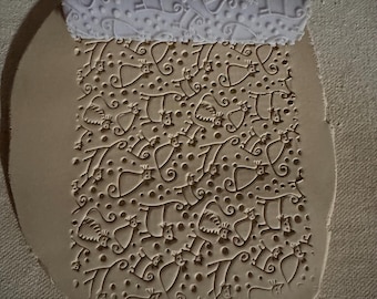 May include: A white rolling pin with a repeating cat design is pressed into a piece of clay, creating a textured pattern. The clay is light brown, and the design includes cats, dots, and swirls. The rolling pin is at the top of the image.