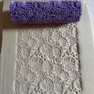 May include: A purple rolling pin with a tropical leaf pattern is positioned above a clay slab with the same embossed design. The rolling pin is cylindrical and features a repeating pattern of monstera leaves and dots. The clay slab shows the impressed design.