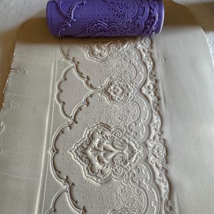May include: A purple patterned rolling pin is pressed into a light-colored clay, creating an embossed design. The embossed pattern features a repeating floral and scrollwork motif, ideal for crafts and decorative projects.
