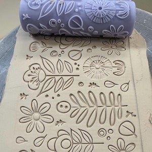 May include: A lavender clay roller with a floral design is pressed into a slab of clay, creating an embossed pattern. The design includes flowers, leaves, and other botanical elements. The roller is approximately 6 inches long.