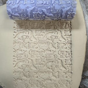 May include: A lavender rolling pin with a Halloween-themed pattern of ghosts, bats, and stars. The rolling pin has a textured surface, and the pattern is imprinted on a light-colored surface, creating a repeating design.