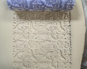 May include: A lavender rolling pin with a Halloween-themed pattern of ghosts, bats, and stars. The rolling pin has a textured surface, and the pattern is imprinted on a light-colored surface, creating a repeating design.