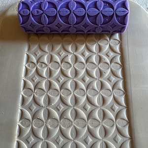 May include: A purple patterned rolling pin next to a piece of clay with an embossed floral and diamond design. The rolling pin has a repeating pattern of flowers and diamonds. The clay has a similar embossed pattern, creating a textured surface.