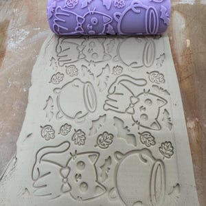 May include: A lavender-colored cylindrical rolling pin with a raised pattern of cats wearing bow ties, cauldrons, and leaves. The rolling pin is shown pressing its design into a flat, light gray slab of clay, creating a repeating embossed texture.