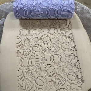 May include: A light purple clay roller with a pumpkin, leaf, and acorn pattern, used to imprint the same design onto a sheet of clay. The clay sheet shows the embossed pattern of pumpkins, leaves, and acorns.