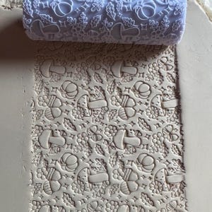 May include: A light purple rolling pin with a repeating pattern of mushrooms, acorns, leaves, and flowers. The rolling pin is pressed into a light brown clay, creating an embossed design. The design is suitable for crafts and baking.