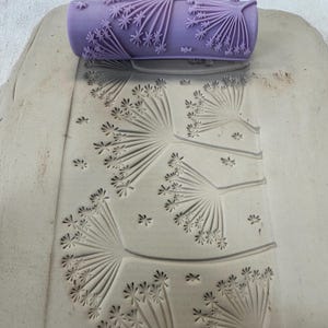 May include: A cylindrical purple clay texture roller featuring a raised dandelion or wildflower pattern. The roller is positioned at the top of a flat, light gray slab of clay, which displays a deeply impressed floral design from the tool.